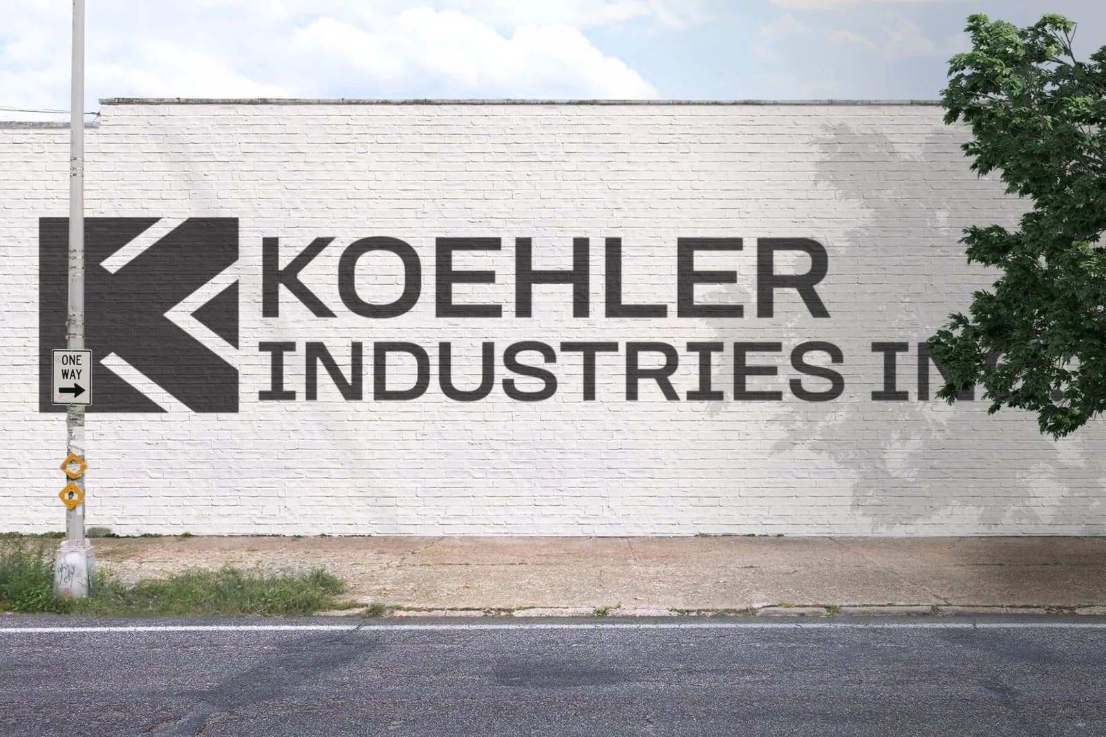 The new Koehler Industries logo — a bold angular K mark and wordmark — painted large in dark gray on a white-painted brick building wall along a street.