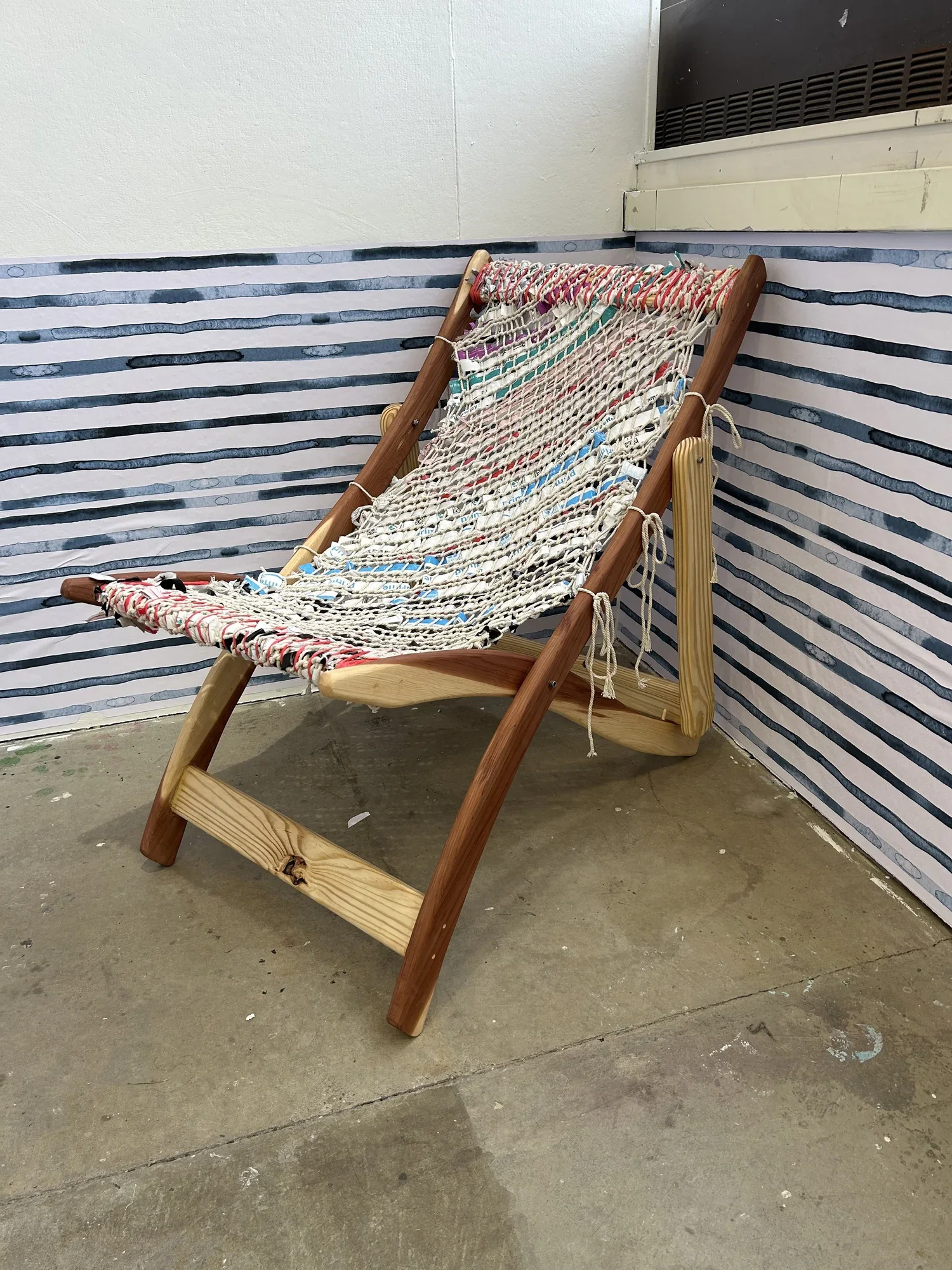 The full-scale finished folding sling chair, its seat woven from collected shoreline plastic waste in red, blue, and white, against a painted blue-stripe water backdrop.