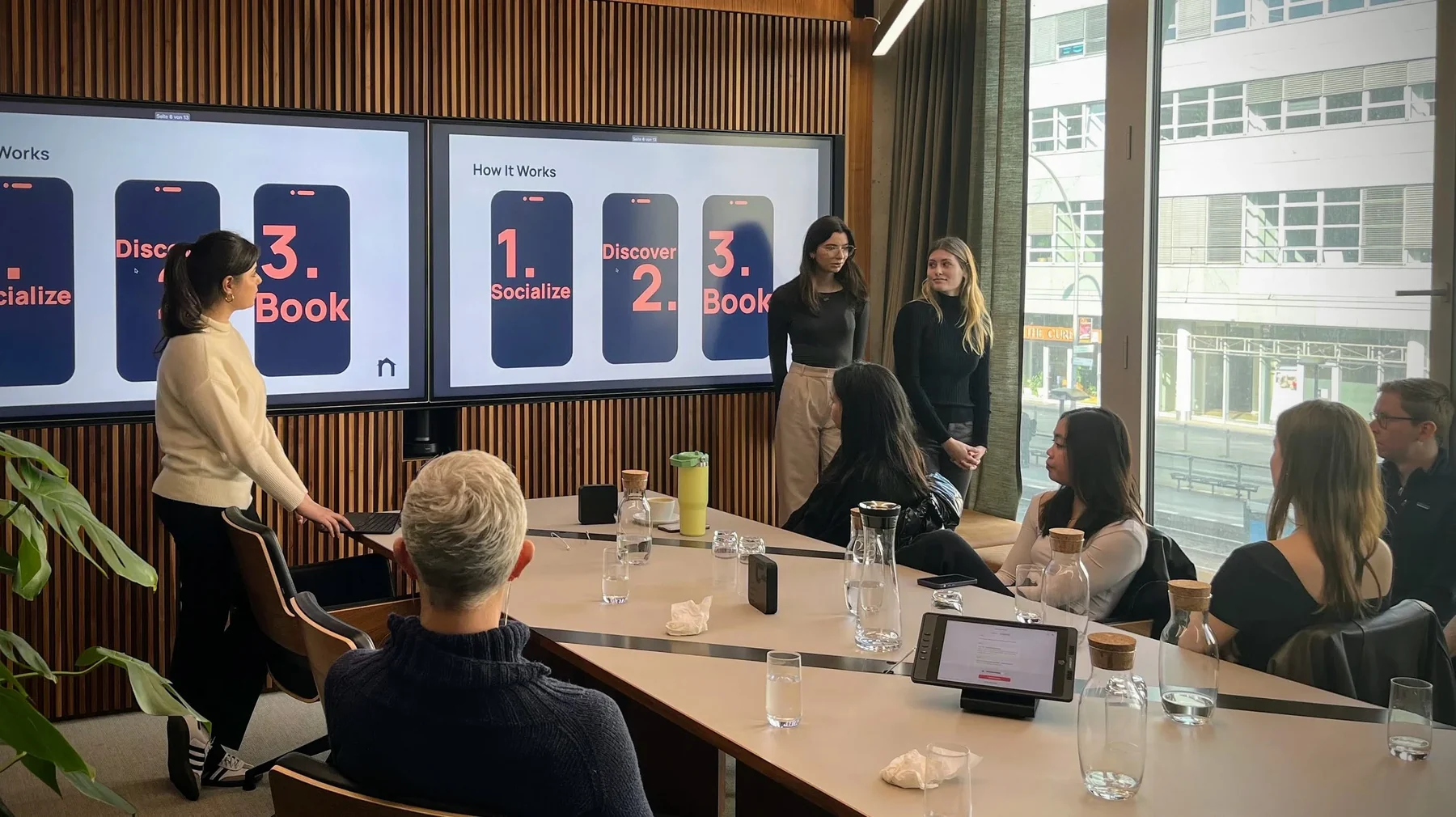 Grace and a teammate presenting the Nomad pitch to a seated audience, with a 'How It Works — Socialize, Discover, Book' slide on the screens behind them.